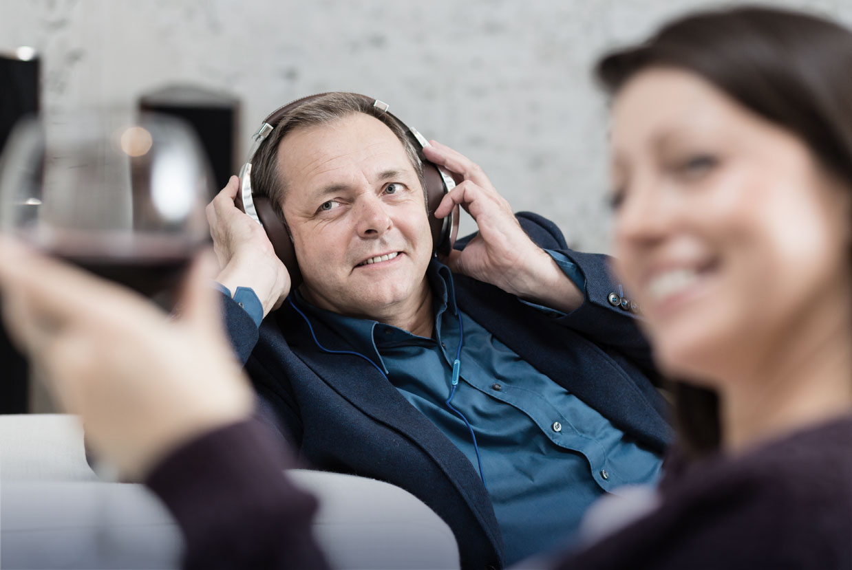 Mann im Hintergrund hört Musik über Kopfhörer. Seine Frau im Vordergrund trinkt Wein.